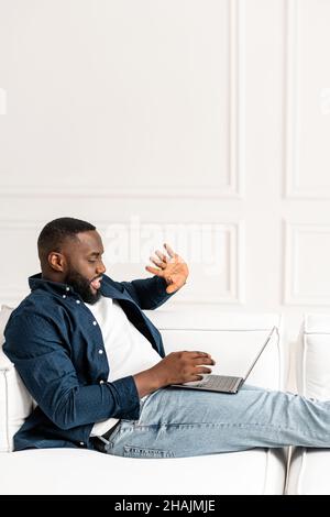 Vertikales Porträt eines erfolgreichen afroamerikanischen Mannes, der per Videoverbindung begrüßt wird, lächelt, zu Hause auf dem Sofa liegt, eine neue Ebene des Arbeitsplatzes, Telekommunikation, mit Laptop auf dem Schoß, Treffen mit dem Team Stockfoto