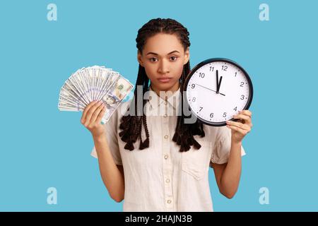Porträt einer ernsten, unzufrieden wirkenden Frau mit schwarzen Dreadlocks, die Dollarnoten und Wanduhr in einem weißen Hemd halten, streng auf die Kamera schauen. Innenaufnahme des Studios isoliert auf blauem Hintergrund. Stockfoto
