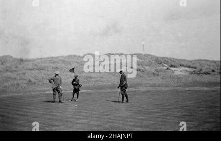 Um 1910, historisch, auf einem Links-Golfplatz, auf einem Putting Green, zwei Golfer in der formellen Kleidung der Ära, einer der in plus zwei, beide mit Jacken, mit einem jungen Caddy, der die Flagge hält, England, Großbritannien. Stockfoto