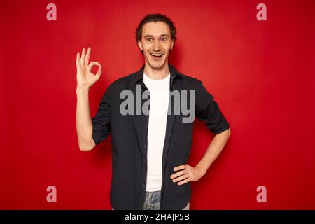 Junger gutaussehender aufgeregt Mann in legerer Kleidung, blaues Hemd, weißes T-Shirt, zeigt ok Zeichen hervorgehoben roten Hintergrund mit Platz für Text, Konzept - erfolgreiche Transaktion, Weihnachtsrabatte, Aktionen. Hochwertige Fotos Stockfoto