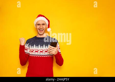 Aufgeregt Mann in Neujahr Pullover, roten Weihnachtshut, isoliert gelben Hintergrund mit freiem Platz für Text der Werbung, mit Telefon in den Händen zeigt Siegeszeichen. Konzept - Gewinn, Verkauf, Rabatte. Hochwertige Fotos Stockfoto