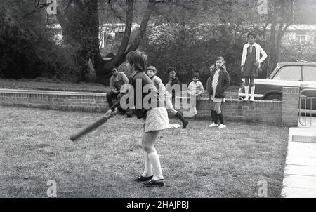 Ende 1960s, historisch, in einem Londoner Vorort, spielen Kinder auf einem Stück Gras draußen an der Straße, ein junges Mädchen schwingt eine Cricket-Schläger, mit anderen Kindern sitzen auf einer niedrigen Wand beobachten, England, Großbritannien. Stockfoto