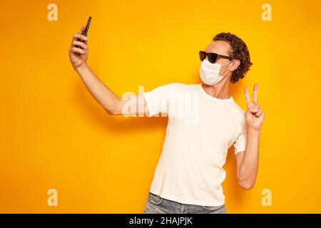 Der Mann nimmt Selfie, macht Videoanruf, zeigt auf die Kamera des Smartphones, genießt Freizeit, bekleidet in legerer Kleidung und Sonnenbrille, zeigt die medizinische Maske friedliche Geste, isoliert auf gelbem Hintergrund. Hochwertige Fotos Stockfoto