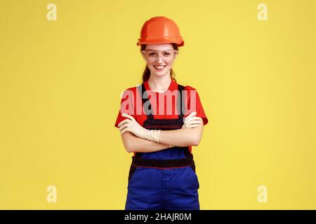 Porträt einer positiven Arbeiterin, die mit gekreuzten Armen steht und selbstbewusst auf die Kamera blickt, Overalls und Helm trägt. Innenaufnahme des Studios isoliert auf gelbem Hintergrund. Stockfoto