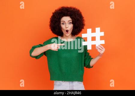 Schockierte Frau mit Afro-Frisur in einem grünen Pullover im lässigen Stil, der auf das weiße Hashtag-Symbol zeigt und den Mund offen hält. Innenaufnahme des Studios isoliert auf orangefarbenem Hintergrund. Stockfoto
