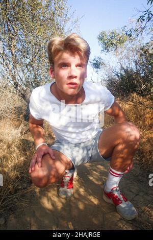 Männliches versteckt sich im Schatten. Jugendliches versteckt sich im Busch. Junger Mann spione auf jemanden. Guckender tom versteckt sich im Busch. Mann im Schatten. Mann in Turnschuhen. Blondes Männchen versteckt sich weg. Stockfoto