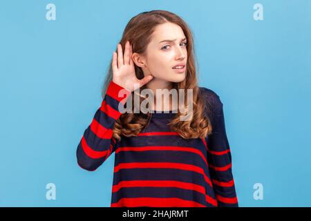 Frau in einem gestreiften Pullover, die Hand am Ohr hält und aufmerksam zuhört, Hörprobleme, Taubheit und Missverständnisse in der Kommunikation hat. Innenaufnahme des Studios isoliert auf blauem Hintergrund. Stockfoto