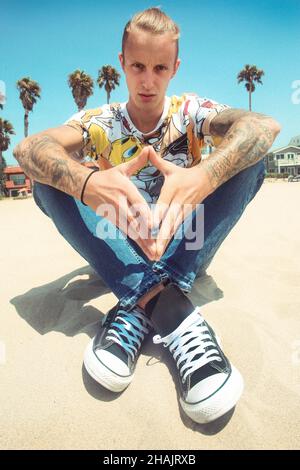 Junger Mann sitzt am Strand. Mann am Strand. Mann hängt am Strand. Nachdenklicher Mann am Strand. Junge am Strand. Teenager entspannt sich am Strandsand. Kerl sitzt. Stockfoto