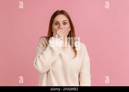 Porträt einer schockierten blonden Frau, die Hand auf den Mund legte und mit Angst in die Augen schaute, schreckliches Geheimnis behielt, trug weißen Pullover. Innenaufnahme des Studios isoliert auf rosa Hintergrund. Stockfoto