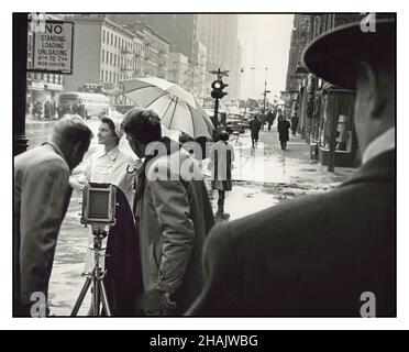 Vintage 1950s Fashion Photography zeigt ein weibliches Model, das im Winter an einer Straßenecke in New York fotografiert wird. Zwei Männer, einer mit einem Regenschirm, stehen hinter der 5x4-Folienkamera, die auf einem Stativ steht. Ein dritter Mann steht im Vordergrund. Mitwirkende Namen Rizzuto, Angelo, 1906-1967, Fotograf 01/1954 [Januar 1954] - Frauen--New York (Bundesstaat)--New York--1950-1960 - Fotografie--New York (Bundesstaat)--New York--1950-1960 - Vereinigte Staaten--New York (Bundesstaat)--New York Gelatin Silberdrucke--1950-1960. Stockfoto