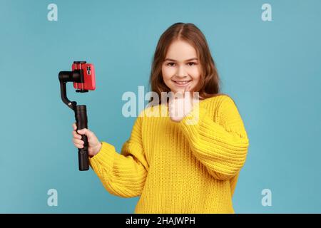 Porträt eines kleinen Bloggers, das Standicam und Handy hält und den Daumen vor die Kamera zeigt, in einem gelben Pullover im lässigen Stil. Innenaufnahme des Studios isoliert auf blauem Hintergrund. Stockfoto