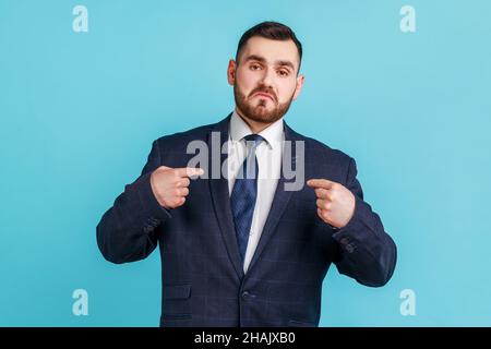 Das bin ich. Porträt eines egoistischen arroganten egoistischen Mannes mit Bart, der einen offiziellen Anzug trägt, der auf sich zeigt und sich mit Erfolg rühmt. Innenaufnahme des Studios isoliert auf blauem Hintergrund. Stockfoto