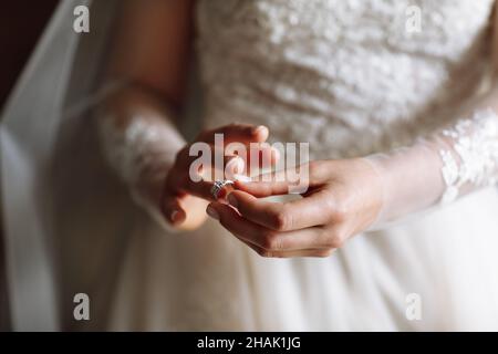 Zugeschnittenes Foto von sanften Händen der Braut, die in einem weißen Spitzenkleid steht, setzt einen Ehering auf ihren Finger. Hochzeitstag. Stockfoto