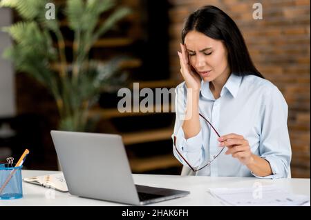 Erschöpfte, überarbeitete kaukasische Geschäftsfrau, Managerin, Sekretärin, müde von der Arbeit mit einem Laptop, sitzend an ihrem Arbeitsplatz, massiert ihren Tempel, hält eine Brille in der Hand und leidet an Migräne Stockfoto