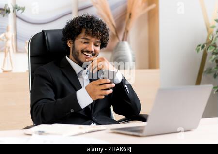 Erfolgreicher, fröhlicher junger indischer Geschäftsmann, Vermarkter oder Manager, der im Büro am Tisch sitzt, einen stylischen Anzug trägt, Smartphone benutzt, Video oder Nachrichten anschaut, mit seinen Kollegen schreibt, lächelt Stockfoto