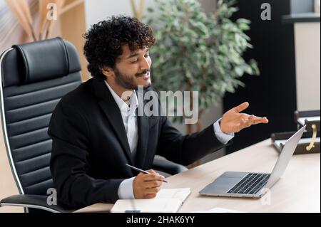 Freundlich glücklich junger Erwachsener smart erfolgreicher indischer Geschäftsmann oder Top-Manager in stilvollem Anzug sitzt am Schreibtisch, spricht per Online-Kommunikation mit Kollegen oder Partnern, Gesten mit der Hand, lächelt Stockfoto
