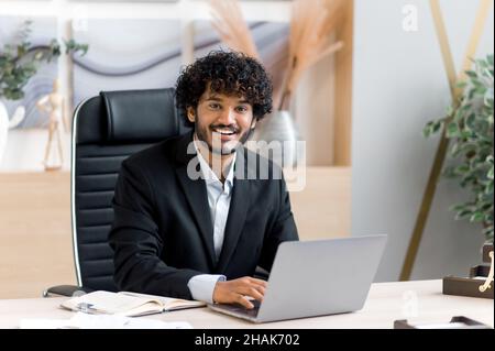 Fröhlicher junger Erwachsener erfolgreicher attraktiver, lockiger indischer Top-Manager, CEO oder Geschäftsmann in stilvollem Anzug sitzt an einem Tisch in einem modernen Büro, nutzt einen Laptop, blickt auf die Kamera und lächelt Stockfoto