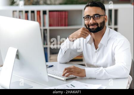 Portrait erfolgreicher smarter glücklicher, gutaussehender indischer Geschäftsmann, Freiberufler oder Manager mit Bart, trägt weißes Hemd, sitzt am Tisch mit Computer im Büro, blickt auf die Kamera und lächelt freundlich Stockfoto