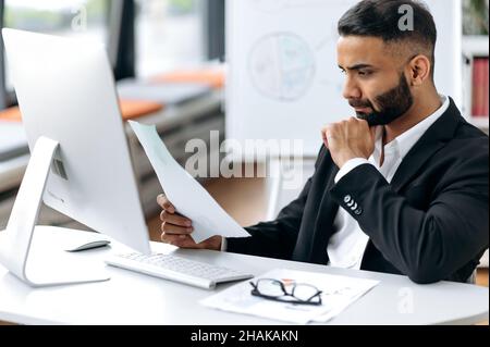 Fokussierter, intelligenter, selbstbewusster indischer Top-Manager oder Geschäftsmann mit Bart, sitzt in formellem Anzug am Tisch, nutzt Computer, durchdacht Dokumente und Diagramme, führt Analysen durch Stockfoto