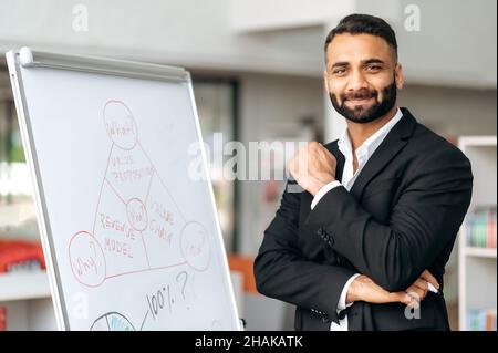 Portrait selbstbewusst erfolgreicher einflussreicher indischer Manager oder Business Coach im formellen Anzug steht mit Diagrammen am Whiteboard, im modernen Büro, blickt auf die Kamera und lächelt freundlich Stockfoto
