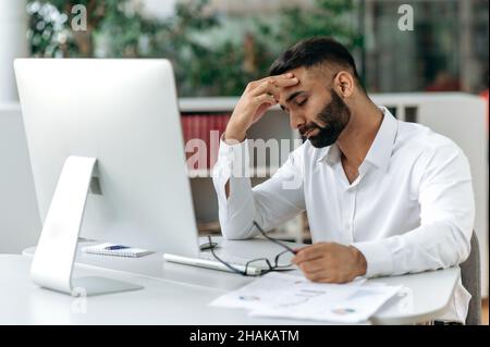 Erschöpfter indischer, junger erwachsener Firmeninhaber, Manager oder Büroangestellter, der mit geschlossenen Augen an seinem Schreibtisch sitzt, seine Stirn reibt, Kopfschmerzen, Stress und Müdigkeit durch Überstundenarbeit empfindet Stockfoto