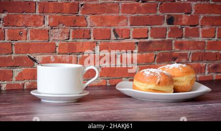 Zwei frische Donuts mit Puderzucker bestreut und eine Tasse Kaffee stehen auf einem Holztisch gegen die Wand. Nahaufnahme. Stockfoto