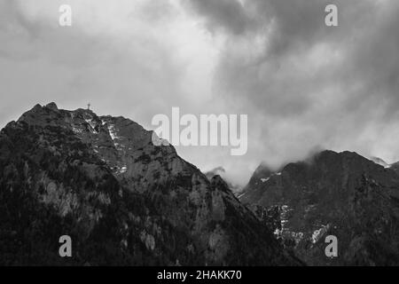 Graustufenaufnahme einer atemberaubenden Wolkenlandschaft über hohen felsigen Bergen an einem düsteren Tag Stockfoto