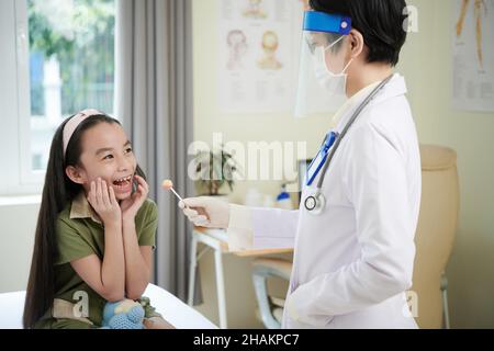 Arzt in schützendem Gesichtsschild und medizinischer Maske, die Lutscher an lächelndes Mädchen aus Angst vor Impfungen gibt Stockfoto