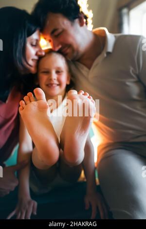 Glückliche Eltern umarmen ihre Tochter, sie sitzt mit ausgestreckten Beinen. Vorderansicht. Stockfoto