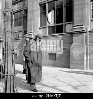 Zwei Pariser Männer in Hüten und Mänteln auf dem Bürgersteig, 1945. Auf einer nicht identifizierten Pariser Straße fängt der Fotograf zwei Männer ein, die wahrscheinlich getrennt voneinander gehen. Die Hauptfigur ist ein älterer Mann mit Bart und Brille, der in tiefer Konzentration zu sein scheint, ohne die Kamera zu kennen. Er verdunkelt teilweise einen jüngeren Mann, der auf den Fotografen zurückblickt. Ein dritter Fußgänger ist meist außer Sichtfeld hinter einem Straßenbaum mit einem aufwändigen Metallkragen. Das Gebäude hinter ihnen hat ein kaputtes Fenster und wurde als Defense d-afficher oder Post No Bill gekennzeichnet. Stockfoto