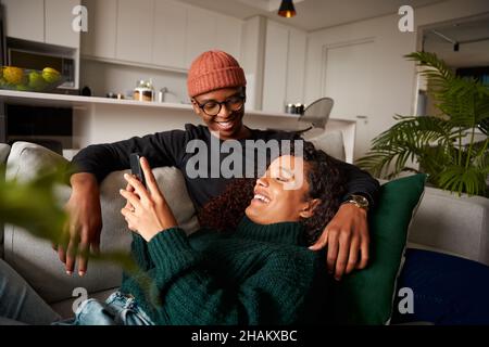 Glückliches multiethnisches Paar, das über Text am Telefon lacht. Auf Sofa in moderner Wohnung Stockfoto
