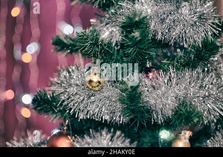 Die kleine gelbe Kugel auf dem Weihnachtsbaum in glänzendem Lametta mit Girlande auf verschwommenem Hintergrund Stockfoto