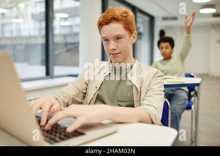 Porträt eines rothaarigen Jungen im Teenageralter, der einen Laptop in einem Codierungskurs für Kinder benutzt, Kopierraum Stockfoto