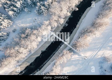 Blick aus der Vogelperspektive auf den Winterwald und den kleinen Fluss mit Brücke. Blick von oben auf den eiskalten Fluss und die schneebedeckte Hängebrücke am sonnigen Wintertag Stockfoto