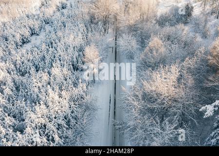 Luftaufnahme von der Drohne der verschneiten Eisstraße an sonnigen Wintertagen. Vogelperspektive Drohne Blick auf leere Asphaltstraße umgeben von schönen Nadelwald. Fliegen Sie über die Winterstraße Stockfoto
