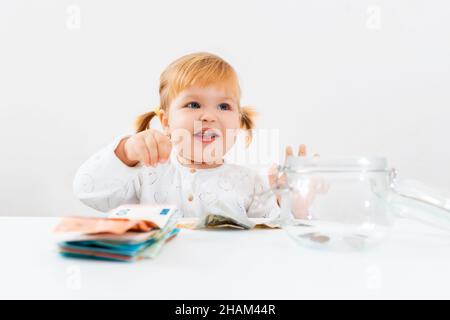 Portrait von kleinen lächelnden niedlichen Mädchen sitzt an einem Tisch mit einem leeren Glas und Stapel Geld. Das Konzept der Finanzierung Bildung und Investitionen. Stockfoto