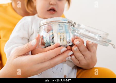 Eine Mutter und ihre Tochter halten zusammen ein Glas mit Geld. Nahaufnahme der Hände. Das Konzept der Finanzierung und Akkumulation von Geldern. Stockfoto
