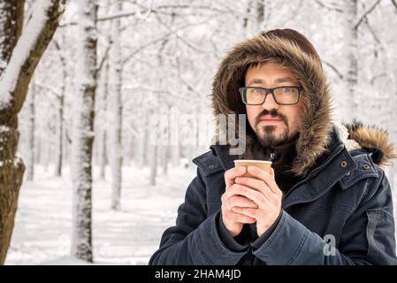 Bärtiger kaukasischer Mann in einer Brille, der Kaffee oder Tee in einer Papiertasse trinkt und am sonnigen kalten Wintermorgen im schneebedeckten Park spazieren geht. Nahaufnahme Porträt eines gutaussehenden Mannes mittleren Alters. Blick auf die Kamera. Stockfoto