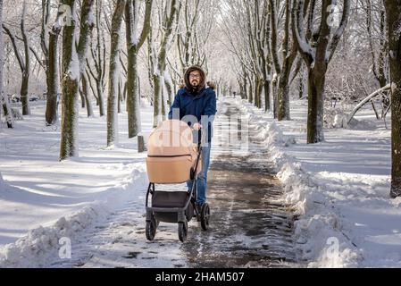 Vater läuft mit beigefarbenem Kinderwagen in einem verschneiten Park. Kalt schöner Wintertag. Entspannende Spaziergänge mit einem Kind. Mann in einer blauen Anorak-Jacke und Jeans. Stockfoto