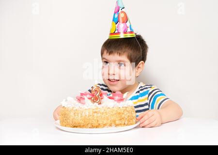 Ein Junge feiert seinen dritten Geburtstag und sitzt mit einem Kuchen an einem Tisch auf weißem Hintergrund und schaut weg. Stockfoto