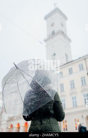 Frau, die im Stadtzentrum von Lviv am Rathaus mit Regenschirm unter Schnee herumläuft. Tourist genießt Architektur im Winter. Reisen in der Ukraine Stockfoto