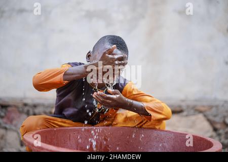 Nahaufnahme eines jungen schwarzen afrikanischen Jungen waschen Gesicht mit frischem, sauberem Wasser aus dem roten Eimer an heißen Tagen Stockfoto