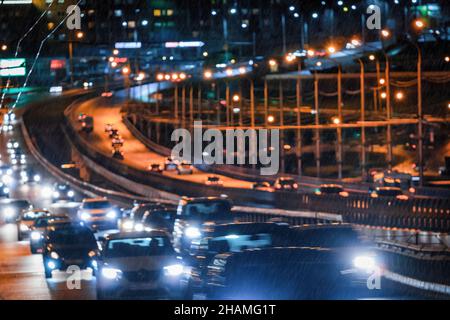 Verschwommene Lichter von sich bewegenden Autos und Laternen, die sich im Regen auf dem nassen Asphalt in der Nachtstadt spiegeln. Konzept für regnerisches Schlechtwetter. Stockfoto