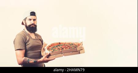 Ein gutaussehender Kurier hält Pappkartons mit Pizza, schlägt vor, leckeres Fast Food zu probieren, trägt Hut und T-Shirt. Stockfoto