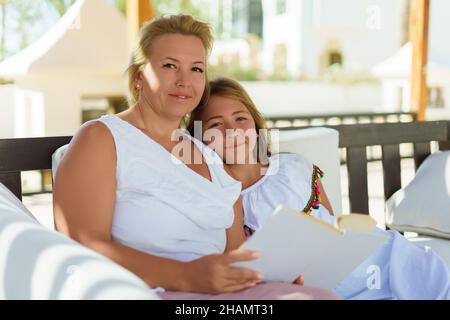 Die kaukasische Mutter mittleren Alters und ihre Teenager-Tochter lesen draußen ein Buch. Das Mädchen legte ihren Kopf auf die Schulter ihrer Mutter, beide blicken auf die Kamera. Selektiver Fokus. Stockfoto