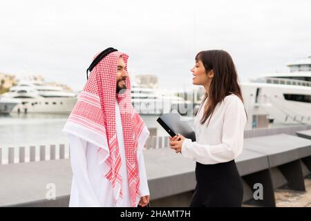 Zwei multirassische Geschäftspartner stehen im Hafen und unterhalten sich nach Abschluss des Deals angenehm, eine junge lächelnde europäische Geschäftsfrau diskutiert mit arabischen männlichen Kunden Vertragsdetails Stockfoto