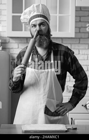 Bärtiger Koch steckt ein Nudelholz in die Nase. Verrückter Koch in einer weißen Küche. Sehr lustiges Foto. Stockfoto