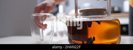 Frau Hand hält transparente Teekannen mit Tee und Glas Tasse Nahaufnahme Stockfoto