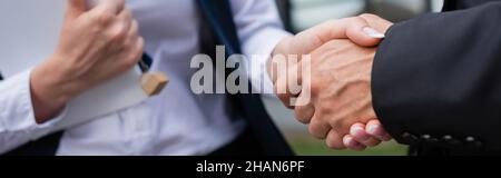 Teilansicht eines Geschäftsmannes, der sich mit verschwommenem makler mit digitalem Tablet und Schlüssel im Freien die Hände schüttelt, Banner Stockfoto