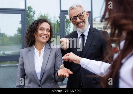 Glückliche afroamerikanische Geschäftsfrau, die Schlüssel von verschwommenem makler in der Nähe aufgeregter Kollege mit Erfolgsgeste nimmt Stockfoto